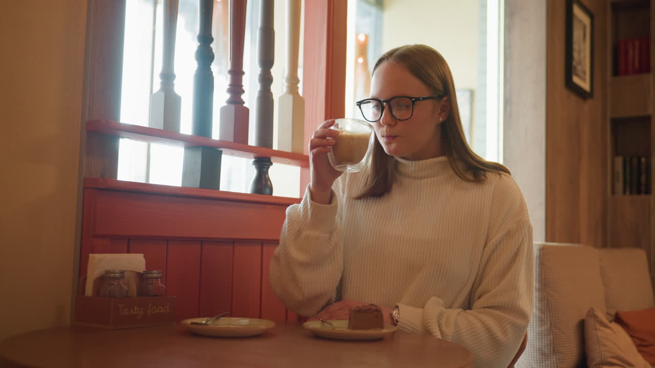 woman in white sweater relaxes at cafe table sipping warm latte with peaceful expression, chocolate dessert on plate, soft lighting from window creates cozy ambiance
