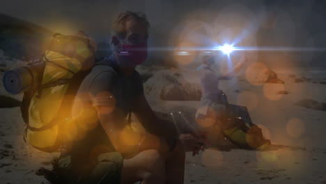 two hikers pausing on beach holding phone and showing backpacks with animated lens flare for health