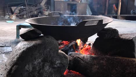 Close-up-of-large-cooking-wok-smoking-over-open-fire-inside-a-basic-kitchen-with-chickens-in-rural-countryside