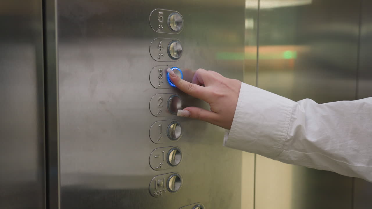 Close up of lady's hand pressing button on stainless steel elevator control panel with floor numbers and braille markings visible, suggesting modern infrastructure inside building interior