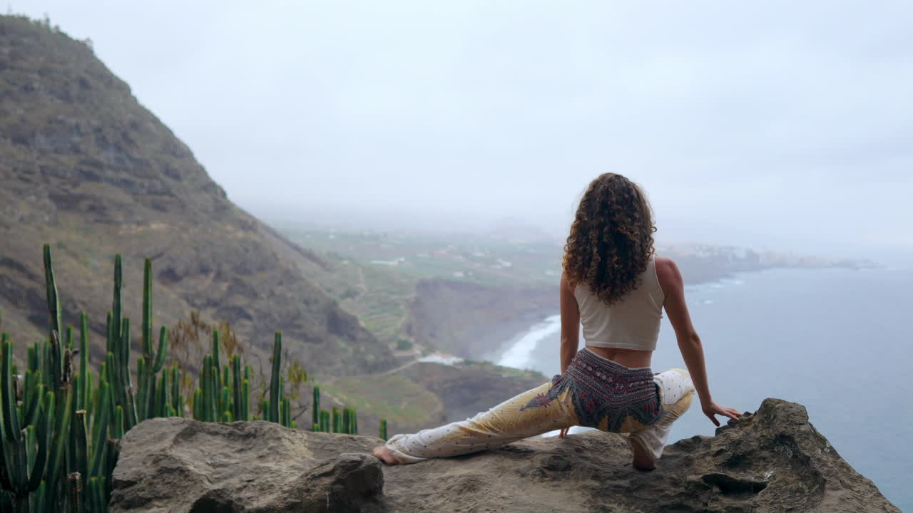 With the ocean spread before her, a woman on a cliff's edge raises her hands in a warrior pose, taking in the sea air during her yoga practice
