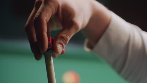 close-up of male hand carefully applying chalk to cue stick before executing precise shot on green pool table. focus on preparation, skill, and accuracy in billiards with blurred balls in background