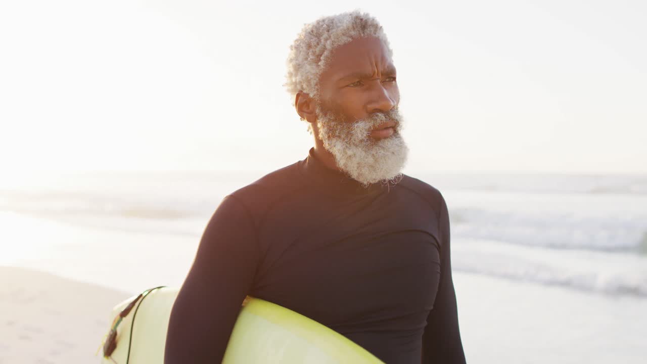 Pria Afrika-Amerika senior yang bahagia berjalan dengan papan selancar di pantai yang cerah.
