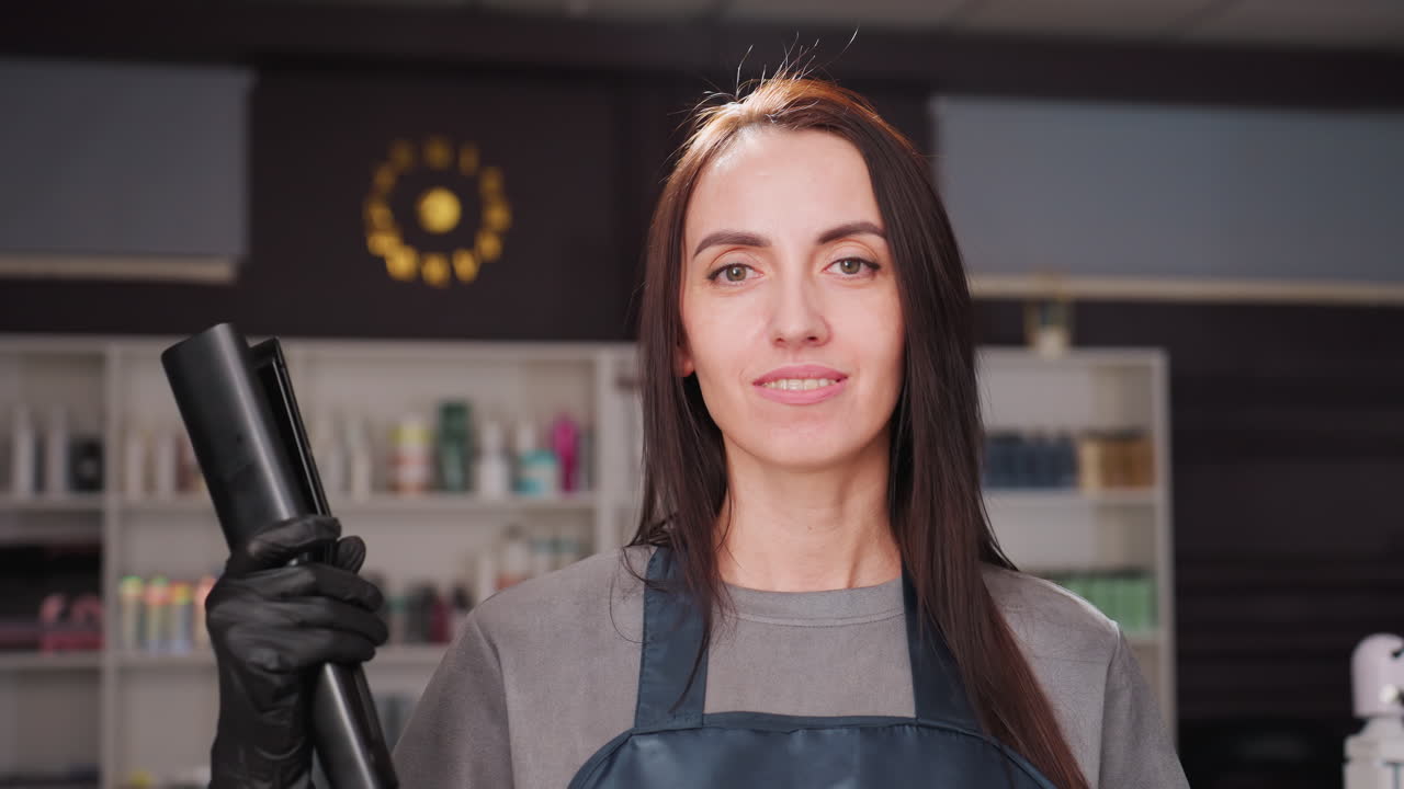 Estilista blanco en un salón sujetando una máquina de cortar; el dueño del estudio posa con seguridad llevando un delantal y guantes; estantes de productos para el pelo desenfocados al fondo; primer plano con mirada directa; herramientas profesionales.
