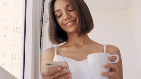 mujer usando el teléfono y bebiendo café junto a la ventana