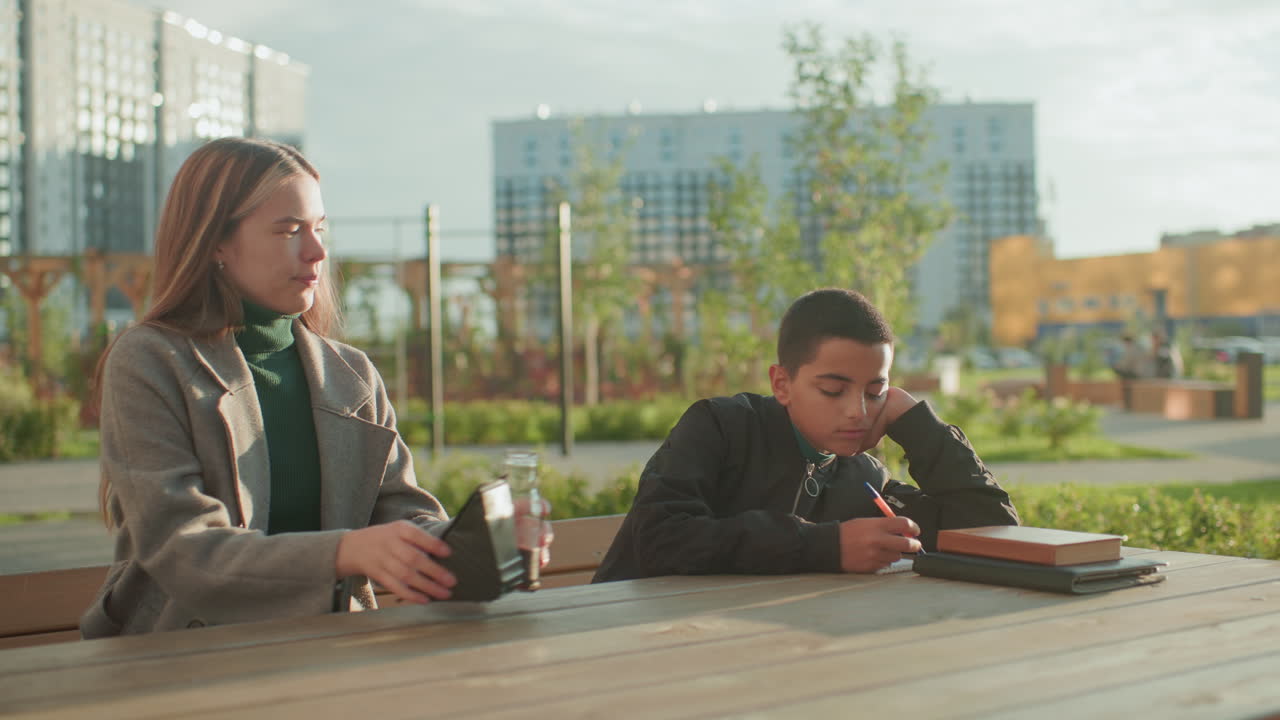 Calm boy writing on wooden table outdoors looks focused then brightens up with happiness as mother offers him snacks, capturing emotional shift and warmth of supportive family care