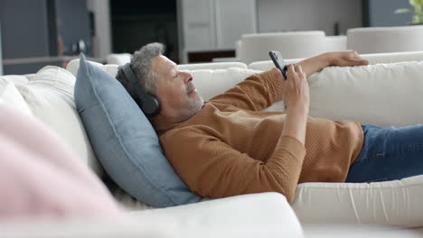 Hombre-Birracial-Mayor-En-El-Sofá-Con-Auriculares-Escuchando-Música-En-Un-Teléfono-Inteligente,-Copiando-Espacio,-Cámara-Lenta