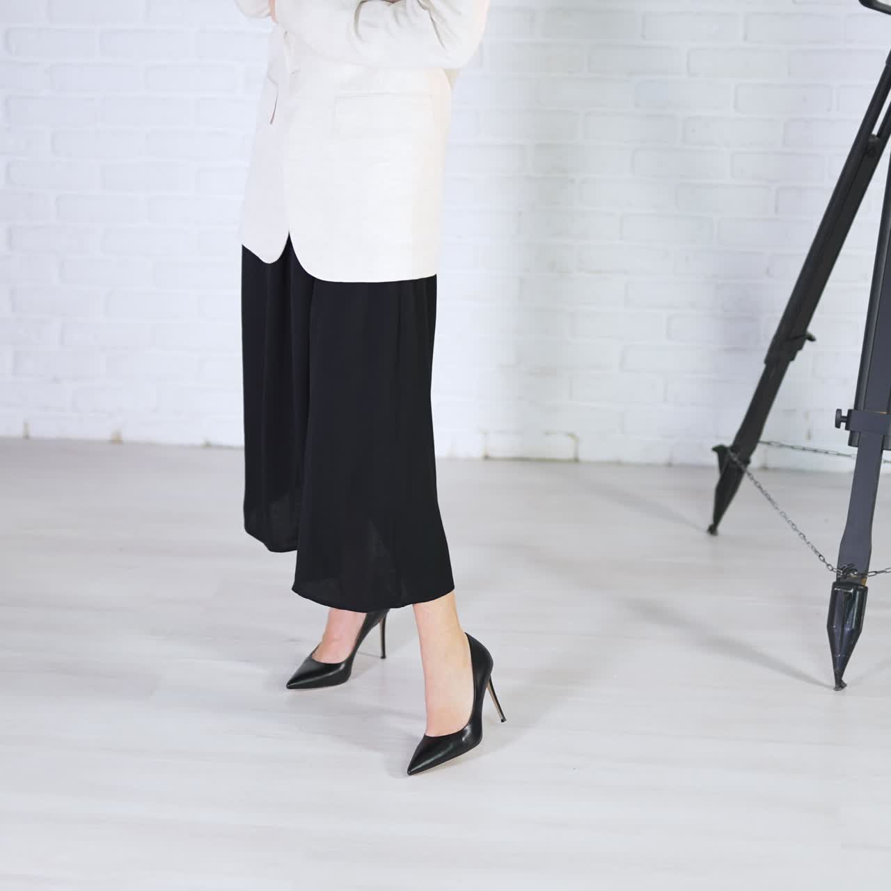Demonstrating black high-heeled shoes in studio. Woman in long dress and white jacket poses in classic shoes. White backdrop