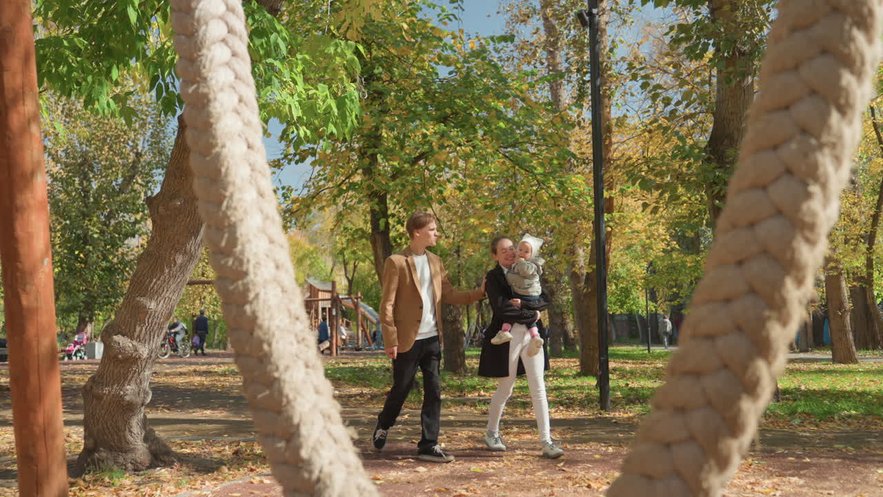 Paseo por el parque de una pareja con un bebé caminando bajo los árboles, luz solar otoñal y un columpio de cuerda trenzada en primer plano, jóvenes padres casados compartiendo una conversación tranquila, miradas tiernas y suave toma de manos.