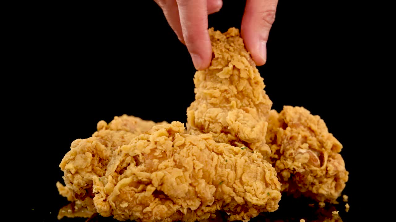 A hand picks up and replaces golden, crumbed fried chicken pieces on a reflective black surface, shot in slow motion with dramatic studio lighting