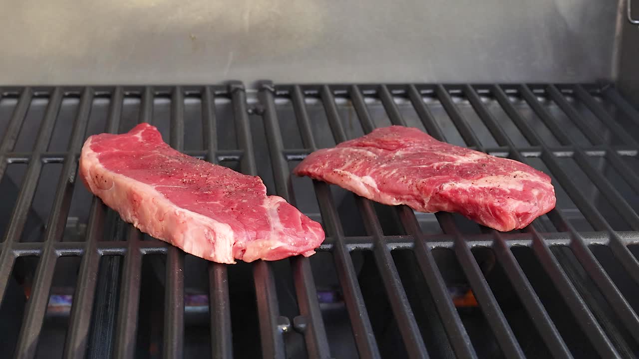 Two beef steaks cooking on a barbecue grill. Close-up view with even lighting and minimal camera movement