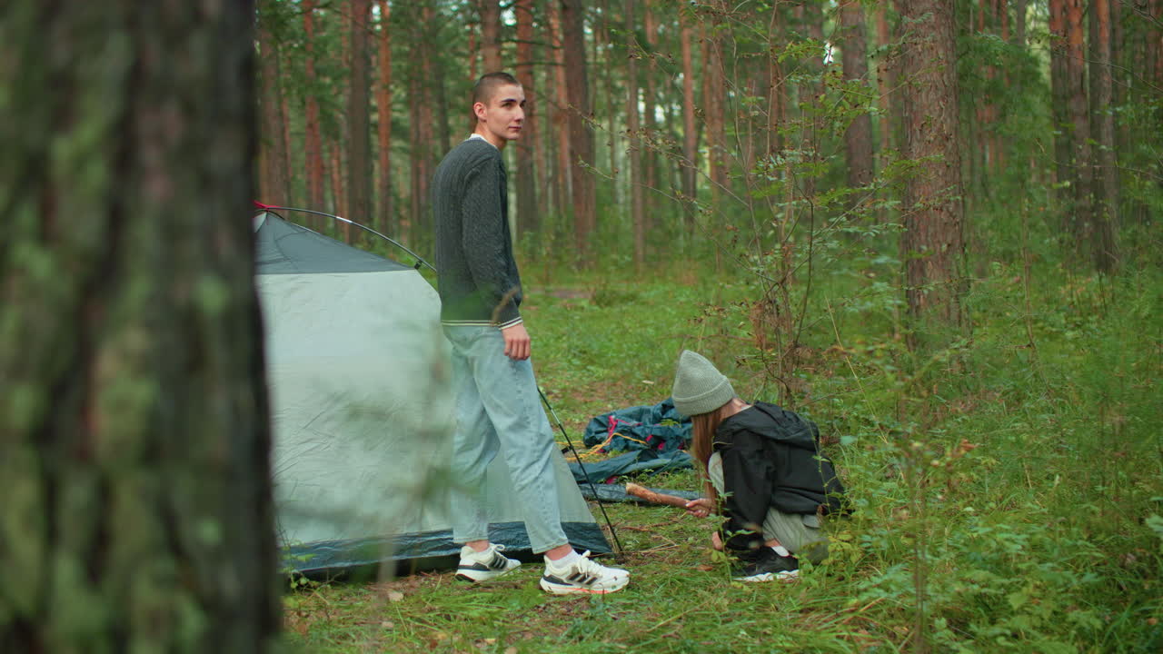 Woman squats near tent in forest while hammering tent peg into ground, briefly lifting head to look at man standing nearby with affectionate gaze during serene camping moment surrounded by tall trees
