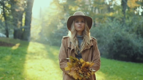 cheerful stylish caucasian young woman wearing a hat and coat having fun in the park, throwing autumn yellow leaves and looking at camera