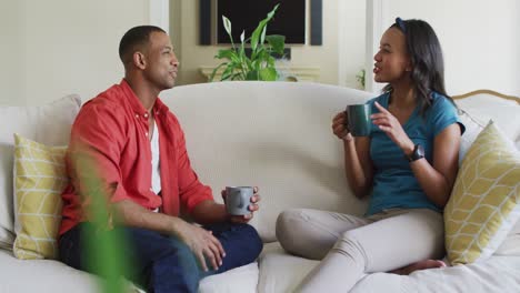 happy biracial couple sitting on sofa with coffee and talking