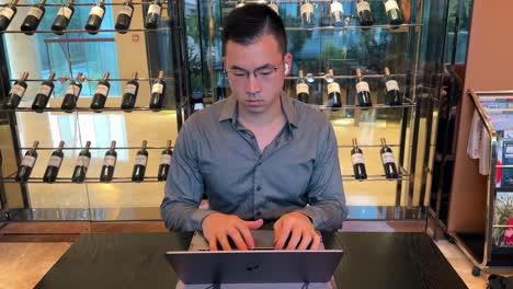 asian millennial entrepreneur man working on laptop inside wealthy luxurious home with wine collection in backgorund