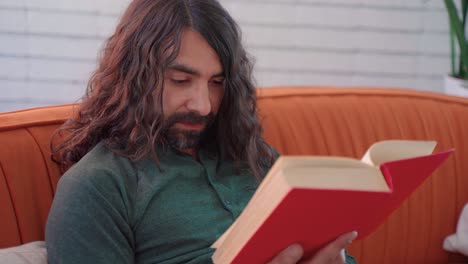 hombre hispano con cabello castaño largo y barba leyendo un libro rojo sentado en un sofá naranja en una habitación blanca
