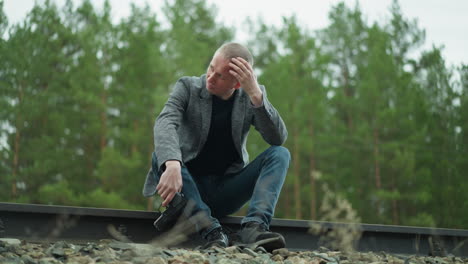 a man in a gray jacket and blue jeans is seated on a railway track, holding a handgun with his head bowed and hand resting on his head, the background features a blurred green forest