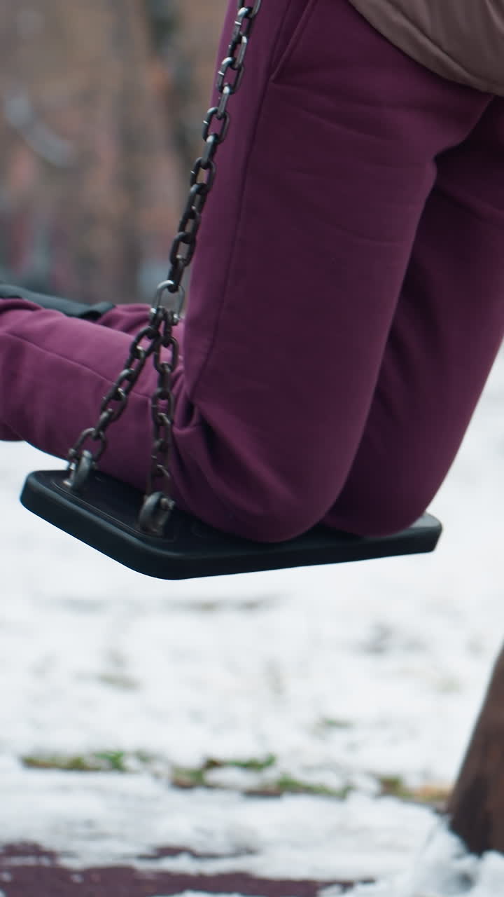 нижний вид тела человека в коричневых брюках и черных сапогах, мягко качающийся, окруженный снегом, размытый фон с голыми деревьями, зданием и проезжающей машиной