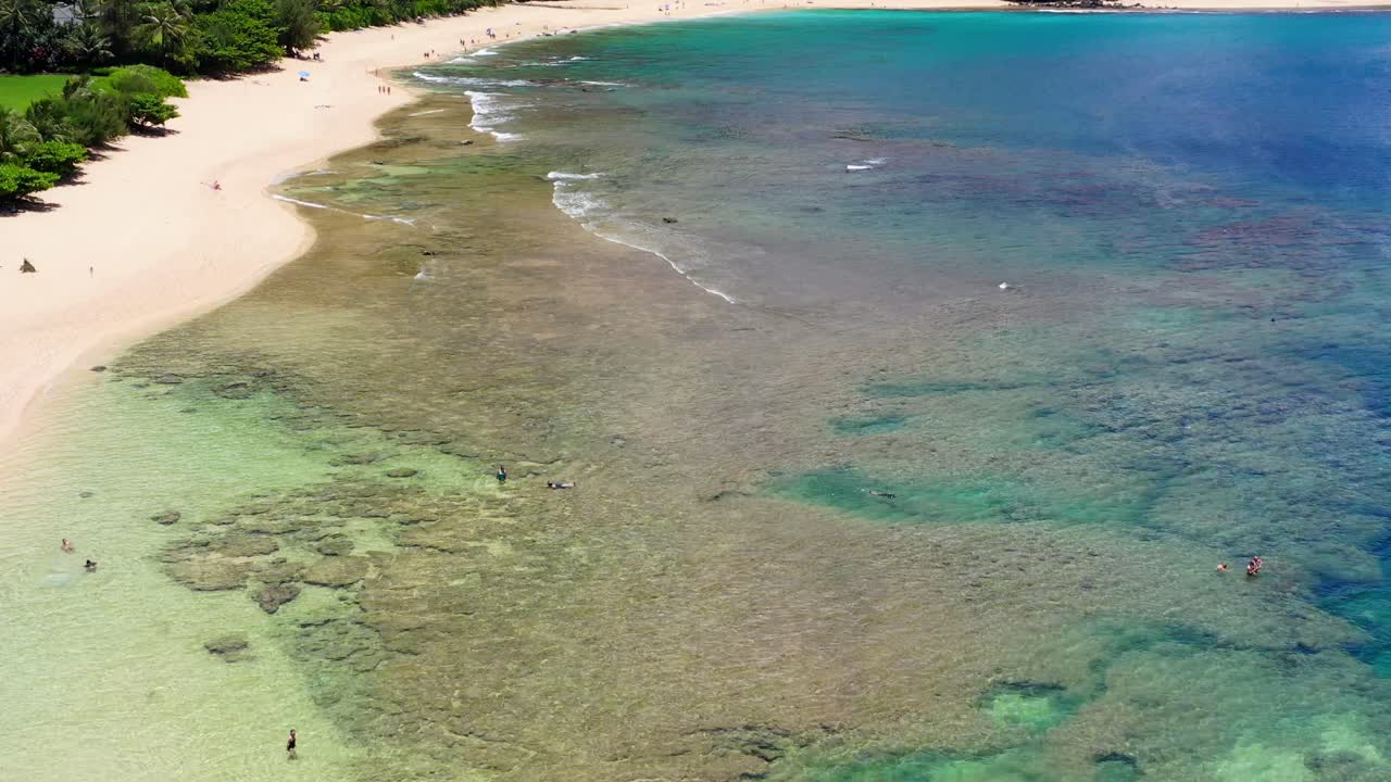 Aerial 4K shot of Tunnels Beach, Kauai. Clear turquoise water, coral reef system, snorkelers, golden sand, and lush mountains—perfect tropical coastline footage for travel or nature content