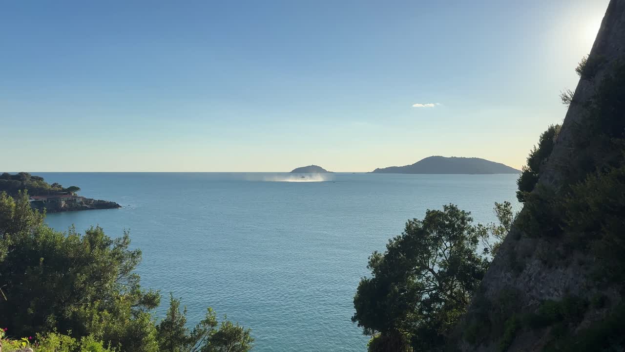 Wide coastal view of the Gulf of La Spezia, Liguria, Italy, with a distant Italian Navy helicopter creating visible spray over water near Tino Island on a clear sunny day