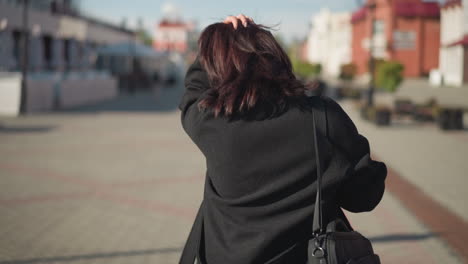 back view of woman adjusting her hair with left hand, carrying black handbag on right arm, walking down paved urban street, background features blurred buildings, greenery, and red pathway