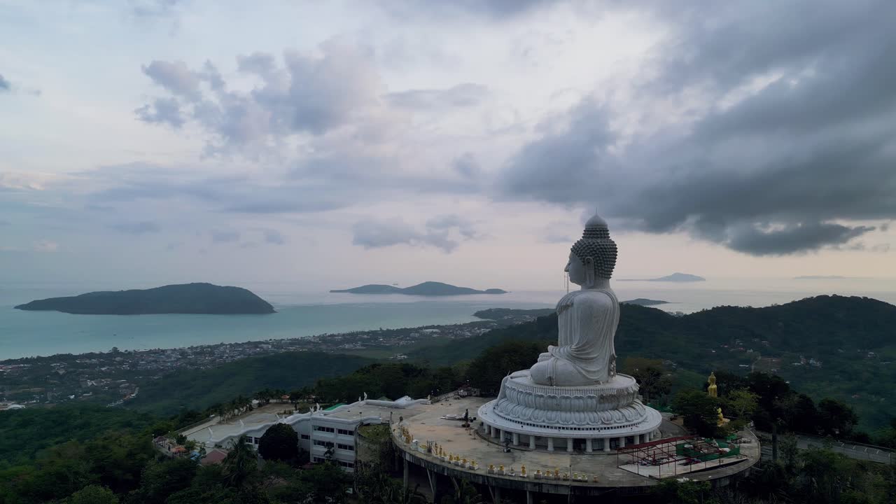 Aerial view of the Big Buddha statue in Phuket, Thailand, with a dramatic cloudy sky and ocean in the background at twilight