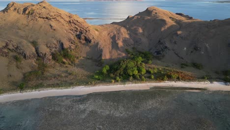 Drone-flying-over-a-coral-reef,-tilting-up-revealing-a-dry-and-barren-island