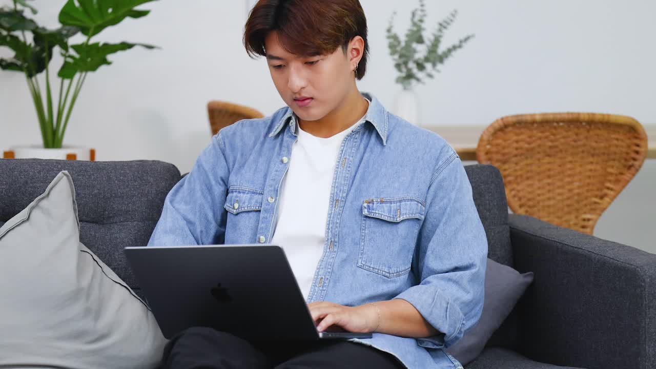 若いアジア人男性が、灰色のリビングルームのソファに座ってノートパソコンで作業している。