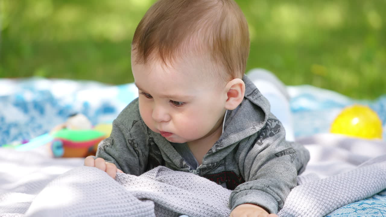 Cute toddler lying on the plaid in the nature. Lovely child looks carefully at the blanket and then pulls it into mouth. Close up.