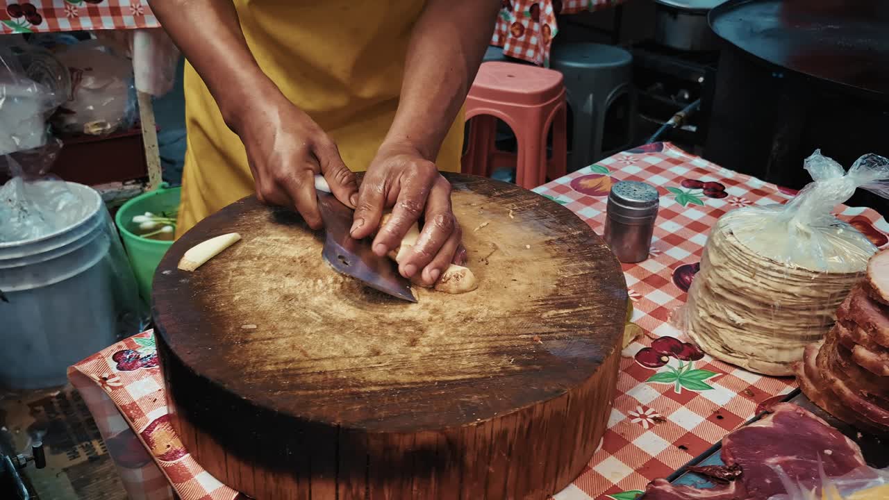 A street food vendor slices a potato with a cleaver on a large round wooden chopping block. Cooking ingredients and utensils are visible on the table