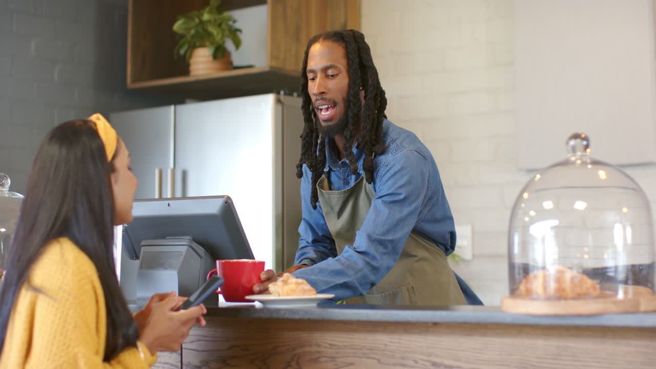 Eine schwarze Barista betritt den Raum, stellt eine Tasse neben das Gebäck an der Theke und grüßt eine indische Frau, die ihr Handy überprüft.