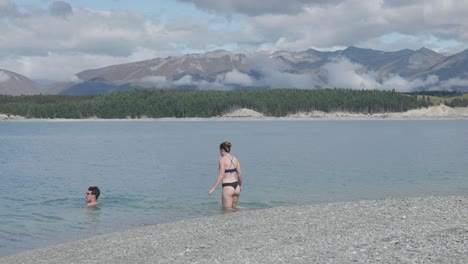 young caucasian couple swimming in a cold blue lake in between forest and mountains on a sunny summer day at lake pukaki, new zealand.