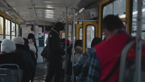 close back view of bundled passengers seated inside moving bus, transparent windows showing bright winter street, soft sunlight and reflections across metal seats create calm urban commute mood