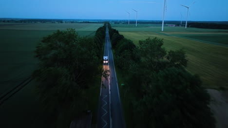 aerial view backwards in front of a rv driving in middle of wind turbines, dusk