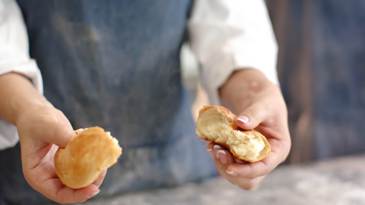 Mid adult woman pinching bun, tearing bun, revealing creamy filling in kitchen wearing denim apron