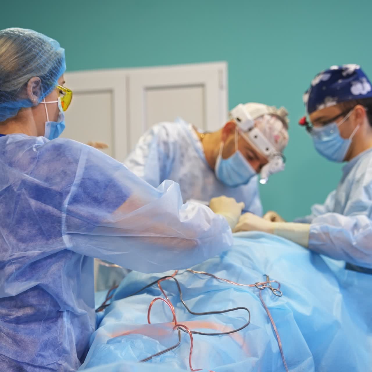 Female nurse in protective suit, cap, mask and glasses arranges instruments for operation. Surgeon operating the patient at backdrop in blur