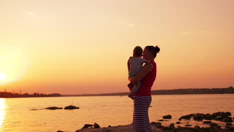 caring and loving mother with her little baby in hands standing near the river. family enjoying time at the nature at sunset.