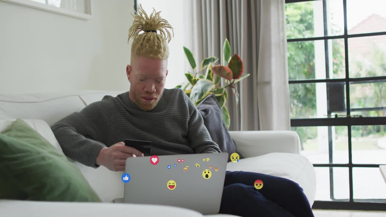 Social media user scrolling phone causing emoji reactions drifting around laptop, smiling at likes