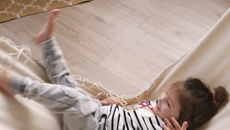 Happy-little-girl-having-fun-indoors,-lying-in-hammock,-and-waving-her-legs.-Smiling-child