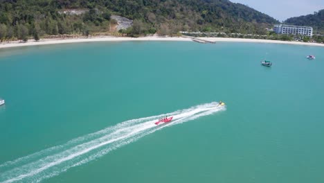 Imágenes-Aéreas-Siguiendo-Un-Barco-Banana-Lleno-De-Gente-Divirtiéndose-Y-Navegando-A-Alta-Velocidad-En-Lancha-Motora,-Deportes-Acuáticos