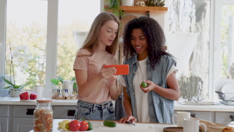 Niña-Negra-Y-Mujer-Joven-Caucásica-Viendo-Un-Video-En-Línea-De-Una-Receta-Vegana-Por-Teléfono-Y-Cocinando.-Dos-Amigas-Hablan-Y-Ríen-En-La-Cocina.-Tiro-Medio.