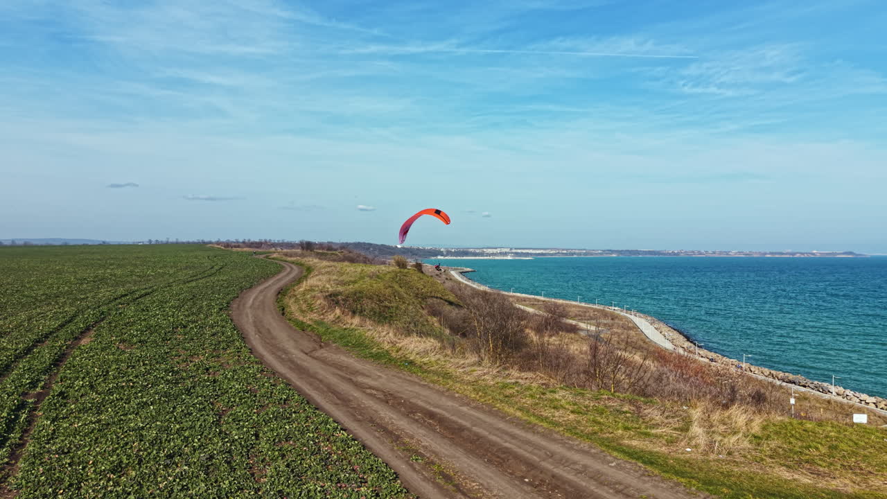 Paraglider flies over coastal field along the shore on a sunny day near the sea with blue sky and distant view of the coastline