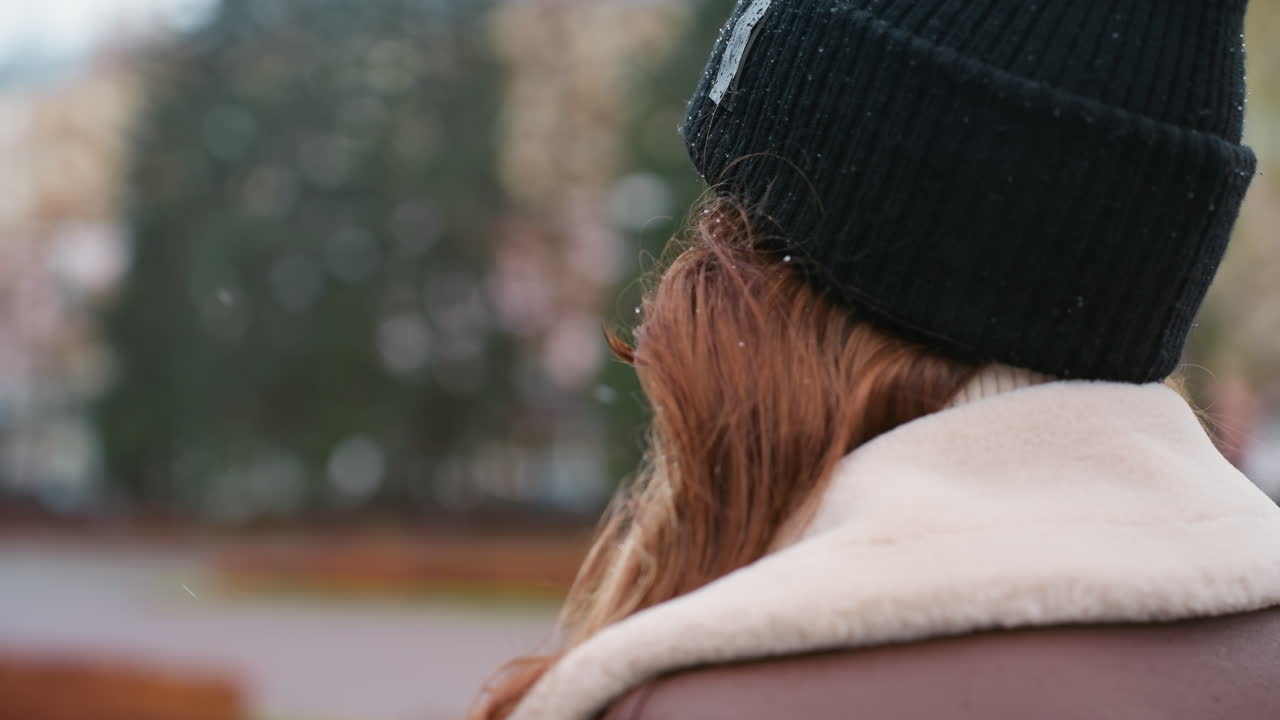 Female in black knit cap and brown shearling jacket walking outdoors on overcast day with blurred autumn colors and soft urban park background