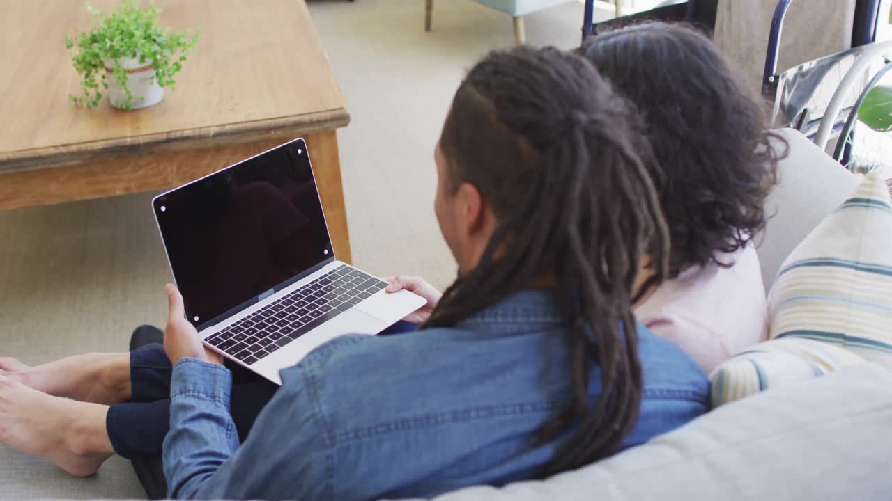 feliz pareja biracial sentada en el sofá usando una computadora portátil en la sala de estar, copia el espacio en la pantalla