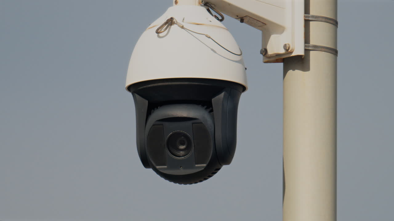Close up of a round, black and white surveillance camera mounted on a white pole with the cloudy sky on the background