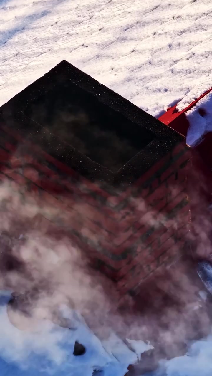 Smoke billows from a red brick chimney on a snowy rooftop in winter.