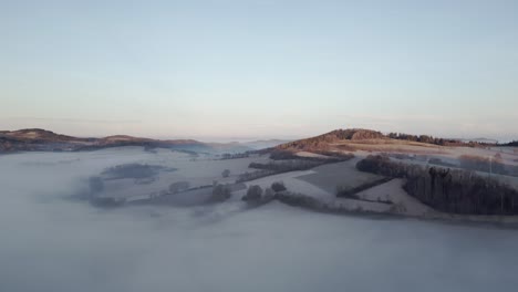 Vista-Aérea-De-Un-Paisaje-Tranquilo-Al-Amanecer,-Con-Colinas-Que-Emergen-De-Una-Espesa-Capa-De-Niebla,-Bañadas-Por-Una-Suave-Luz-Solar.