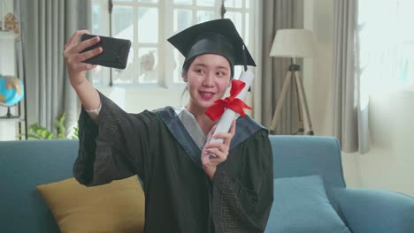 young asian woman taking a selfie while wearing a graduation gown and cap, hand holding university certificate in living room