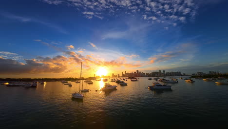 Beautiful scenery of multiple yachts standing on anchor. Amazing sunset over the modern metropolis at backdrop. FPV drone footage.