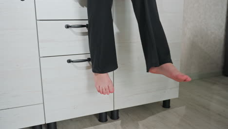 close up of adult legs swinging while seated on white wood cabinet in cozy kitchen interior, black pants draping down, soft light highlighting motion and texture of cabinet surface